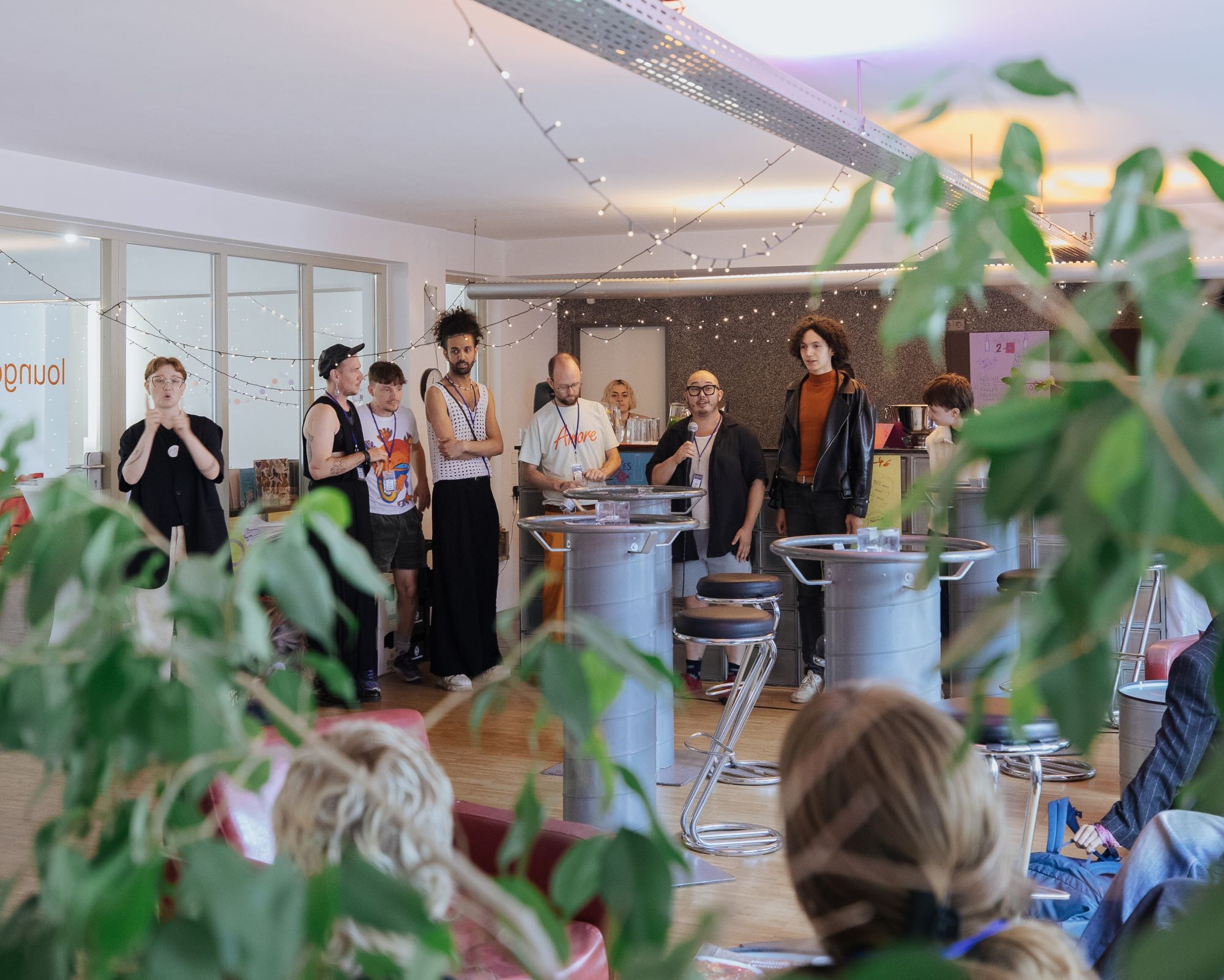 Eight people of the Bonanzafest team stand in front of the bar while addressing the visitors at the community brunch. One of them is translating into sign language.   Acht Personen des Bonanzafest Teams stehen vor der Bar und sprechen zu den Besucher*innen während des Community Brunch. Eine Person des Teams übersetzt simultan in Gebärdensprache. 