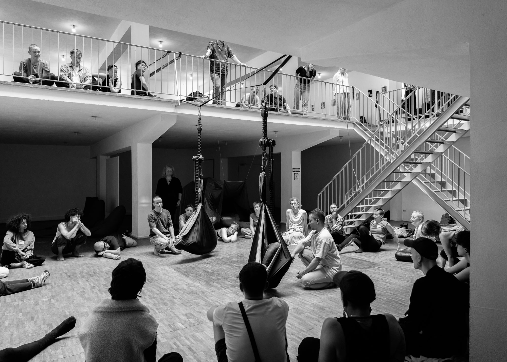 A black and white image of Frederik Marroquín and Dorian Bonelli during their installative performance of ‘Being present in time – 365 days’. The two performers sit on the floor in the middle of the room while looking at two hammocks in front of them, which hang from contraptions from the railing of the gallery level above them. In each hammock is one audience member. The rest of the audience is seated in a circle around the performers.   Eine Schwarz-Weiß-Aufnahme von Frederik Marroquín und Dorian Bonelli während ihrer installativen Performance "Being present in time - 365 days". Die beiden Performer sitzen auf dem Boden in der Mitte des Raumes und blicken auf zwei Hängematten vor ihnen, die an Vorrichtungen am Geländer der Galerieebene über ihnen angebracht sind. In jeder Hängematte sitzt ein Zuschauer. Der Rest des Publikums sitzt im Kreis um die Performer.