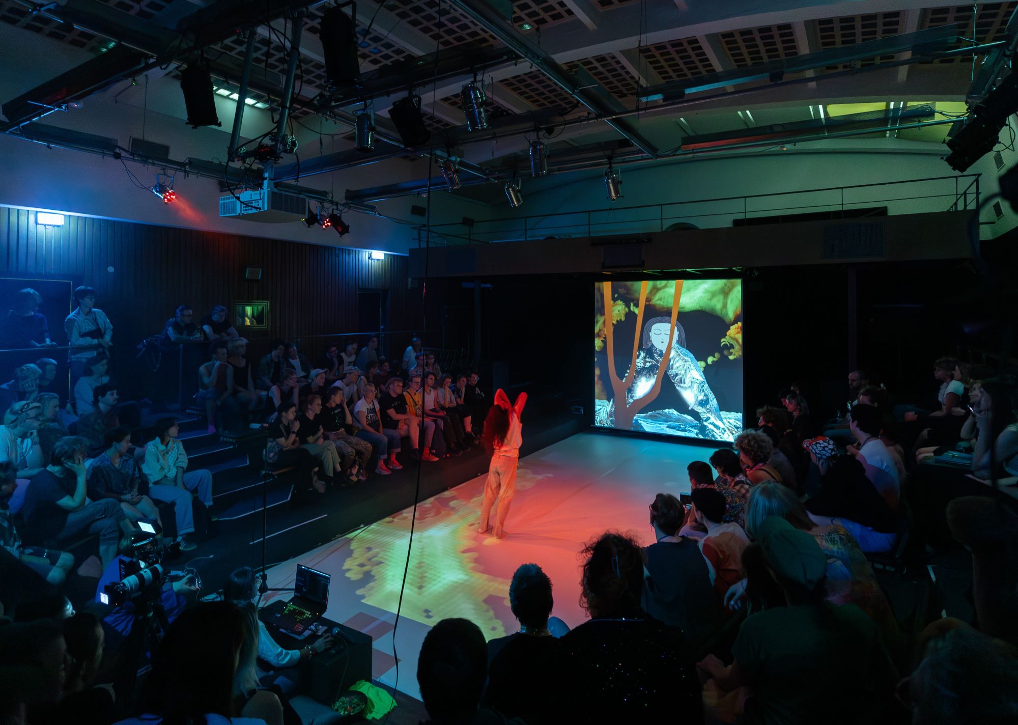 Pachamama, wearing a completely cream outfit, is seen from behind with their long hair falling onto their back while looking upwards. They are standing on a white runway that leads towards a screen at the end of the room, which shows an art installation in green, brown, black and silver tones. Geometric shapes in yellow and orange are projected onto the runway while Pachamama is indirectly lit in red.  The audience is seated in a U-shape around the runway and watches the performance intently.   Pachamama trägt ein cremefarbenes Outfit und ist von hinten und nach oben schauend zu sehen, während deren langes Haar auf deren Rücken fällt. Pachamama steht auf einem weißen Laufsteg, der zu einer Leinwand am Ende des Raumes führt, die eine Kunstinstallation in Grün-, Braun-, Schwarz- und Silbertönen zeigt. Geometrische Formen in Gelb und Orange werden auf den Laufsteg projiziert, während Pachamama indirekt in Rot beleuchtet wird.  Das Publikum sitzt U-förmig um den Laufsteg herum und verfolgt aufmerksam die Aufführung.