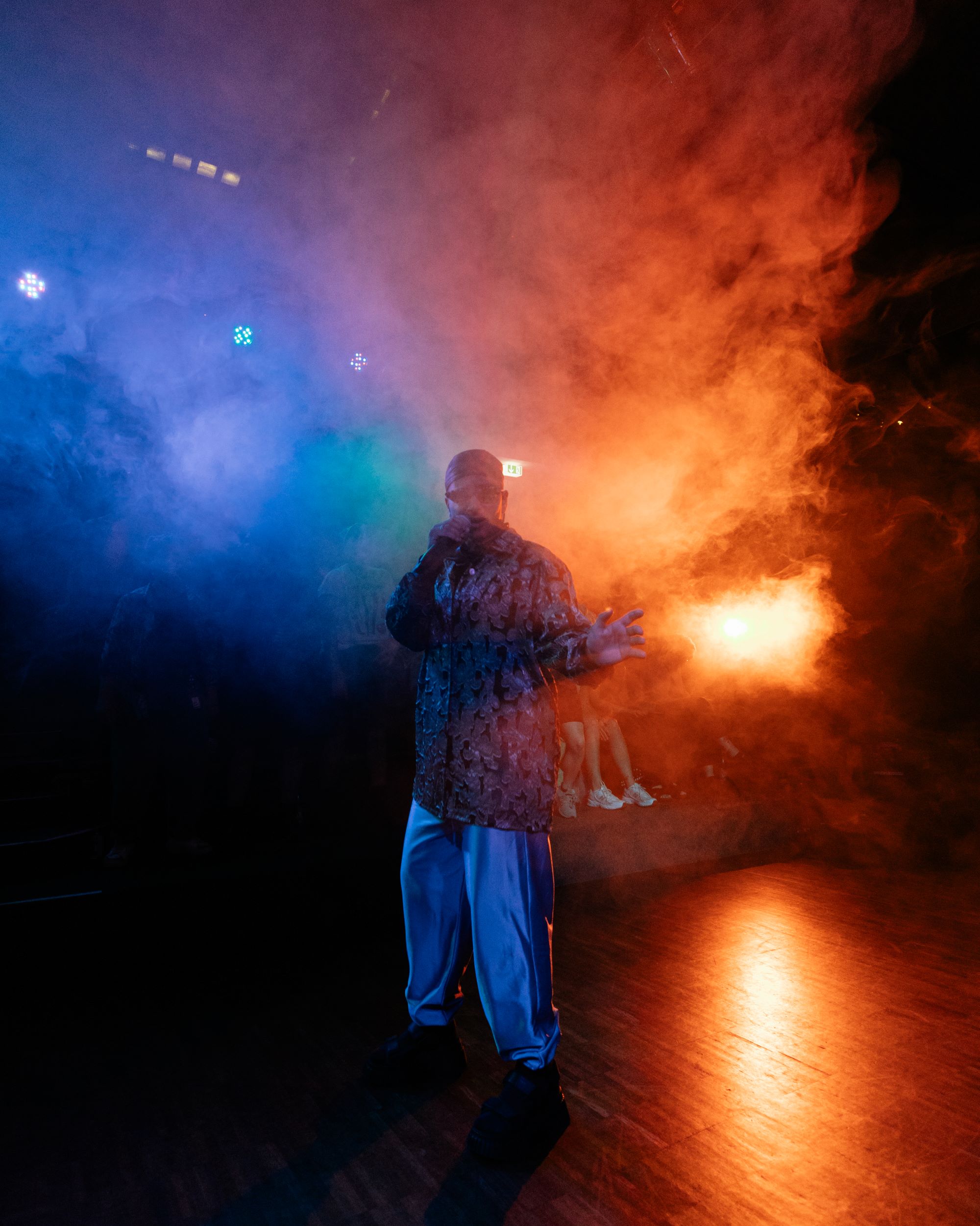 Iranian rapper Säye Skye during their performance. He is looking into the camera while wearing sunglasses, a headscarf, a loose patterned button-down shirt in brown tones, loose silver pants and dark shoes. His right hand is holding the microphone while the left is captured in movement of gesticulation. Säye stands within fog, which fills the room and is illuminated through orange and blue stage lights.   Der iranische Rapper Säye Skye während seines Auftritts. Er schaut in die Kamera, trägt eine Sonnenbrille, ein Kopftuch, ein lockeres gemustertes Button-Down-Hemd in Brauntönen, eine weite silberne Hose und dunkle Schuhe. Seine rechte Hand hält das Mikrofon, die linke ist in einer gestikulierenden Bewegung eingefangen. Säye steht inmitten von Nebel, der den Raum ausfüllt und durch orangefarbene und blaue Scheinwerfer beleuchtet wird. 