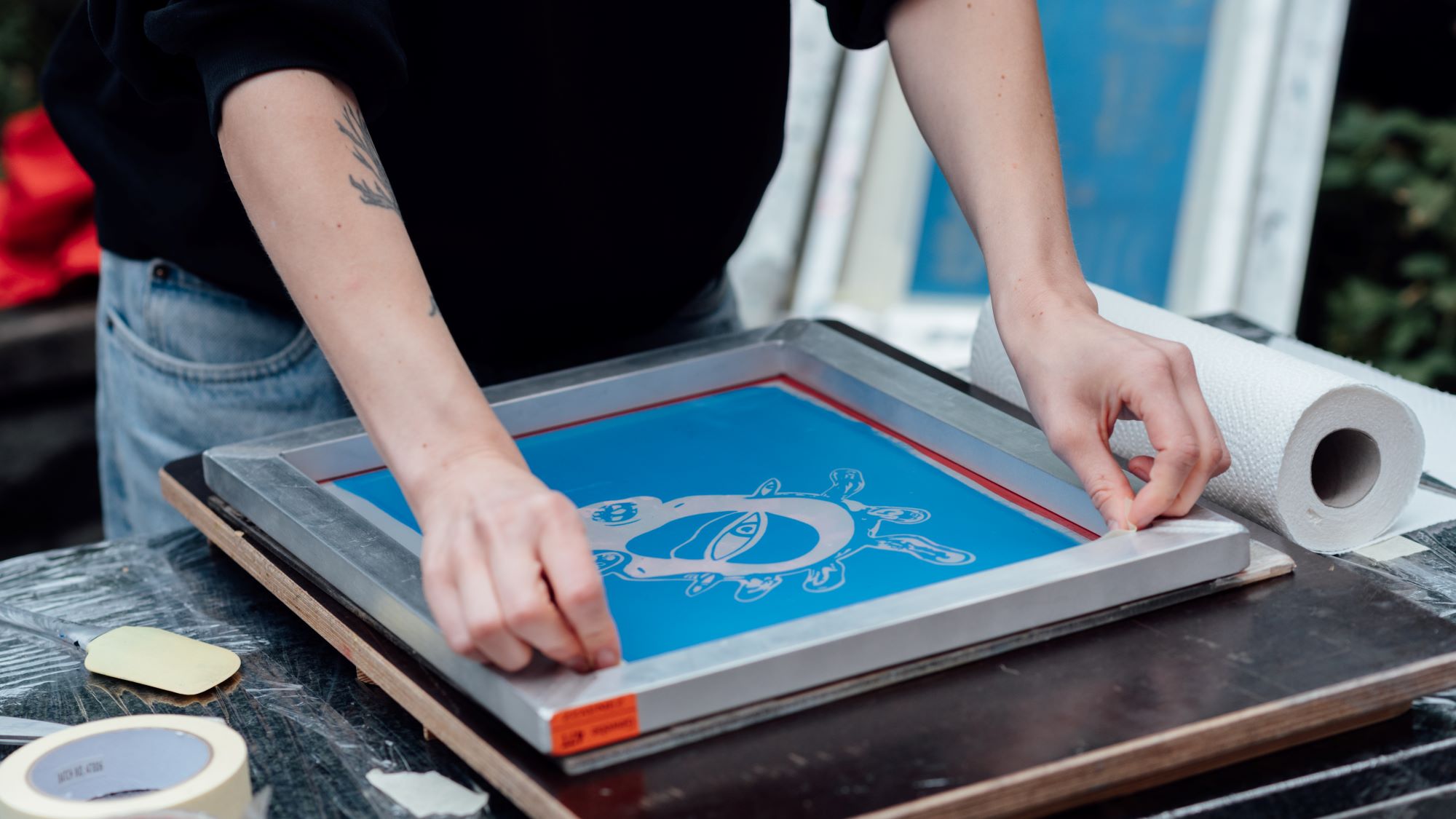 A person in the process of screen printing. The frame only shows their arms as they are holding down the print in front of them on a worktable. Utensils lie next to them on the table.   Eine Person, die einen Siebdruck anfertigt. Der Ausschnitt zeigt nur deren Arme, während die Person den Druck vor sich auf einem Arbeitstisch festhält. Utensilien liegen daneben. 