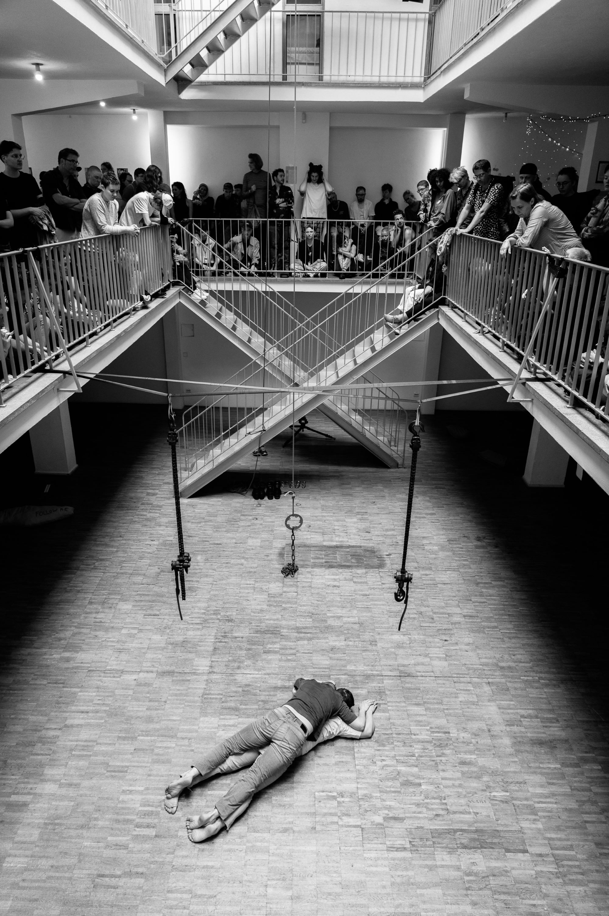 A black and white image of Frederik Marroquín and Dorian Bonelli during their installative performance of ‘Being present in time – 365 days’. The picture is taken from the gallery level of the performance room and shows Frederik and Dorian lying on top of each other on wooden floors and in an empty space. The gallery level is filled with people that stand or sit while watching the performance.   Eine Schwarz-Weiß-Aufnahme von Frederik Marroquín und Dorian Bonelli während ihrer installativen Performance "Being present in time - 365 days". Das Bild wurde von der Galerieebene des Aufführungsraums aus aufgenommen und zeigt Frederik und Dorian, wie sie auf dem Holzboden in einem leeren Raum aufeinander liegen. Die Galerieebene ist mit Menschen gefüllt, die stehend oder sitzend die Performance beobachten.