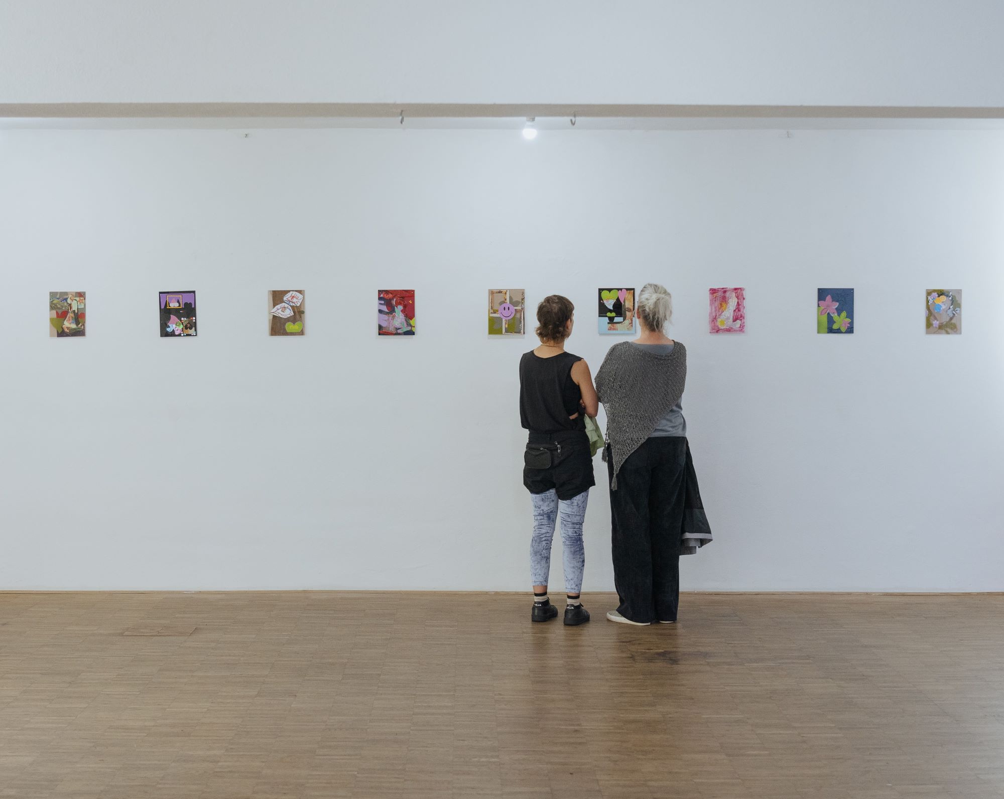 Two visitors stand with their backs turned to the camera and look at a series of colourful artworks that are positioned in a straight horizontal line on a white wall. The visitor on the right is wearing their blonde hair in a ponytail and is dressed in loose black and grey clothing. The visitor on the left has medium length brown hair and is wearing a black tanktop and black shorts over blue and white leggings. They stand close together as if discussing the particular artwork in front of them.   Zwei Besucher*innen stehen mit dem Rücken zur Kamera und betrachten eine Reihe von bunten Kunstwerken, die in einer geraden horizontalen Linie auf einer weißen Wand angeordnet sind. Die Person auf der rechten Seite trägt deren blondes Haar in einem Pferdeschwanz und ist in lockere schwarz-graue Kleidung gekleidet. Die Person auf der linken Seite hat mittellanges braunes Haar und trägt ein schwarzes Tanktop und schwarze Shorts über blau-weißen Leggings. Sie stehen dicht beieinander, als würden sie das Kunstwerk vor ihnen besprechen. 